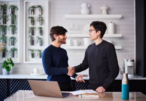 Two people shaking hands beside a desk with a laptop